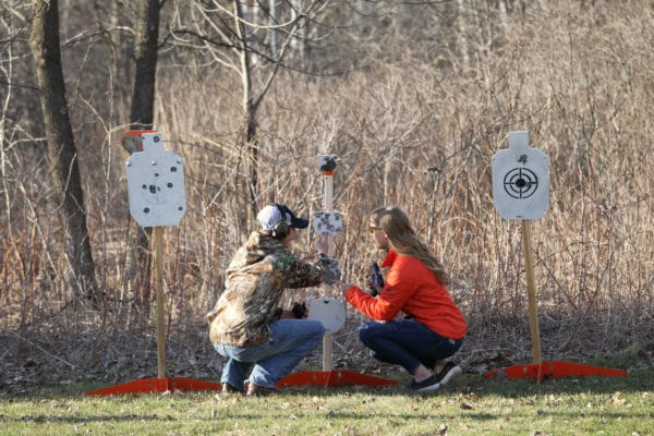 A man and a woman crouch down in front of an array of AR500 steel targets and point at places their gun shots hit the target